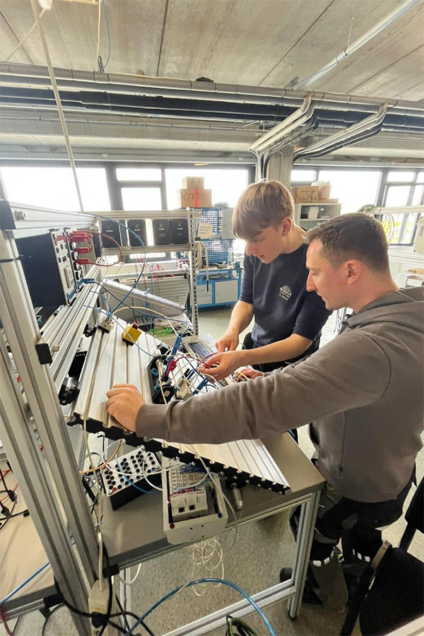 Zwei Personen arbeiten an einer technischen Anlage mit Kabeln und Geräten.